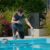 pool-technician-pouring-liquid-into-pool-while-using-tablet-surrounded-by-chemical-containers_0x800